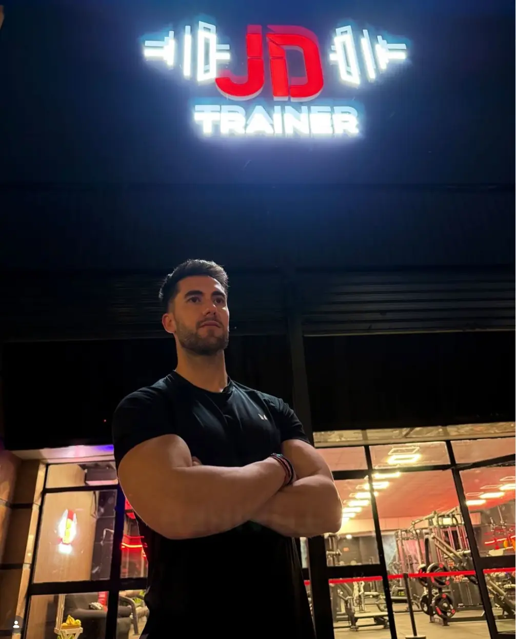 José David, coach de JD Trainer Gym, con los brazos cruzados bajo el letrero luminoso del gimnasio